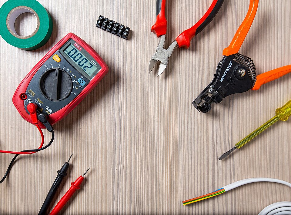 Ein Digitalmultimeter, das eine Messung anzeigt, umgeben von verschiedenen Werkzeugen, darunter eine Zange, eine Abisolierzange, ein Schraubendreher, Isolierband und Kabelverbinder liegen auf einem Arbeitstisch mit strukturierter Oberfläche.