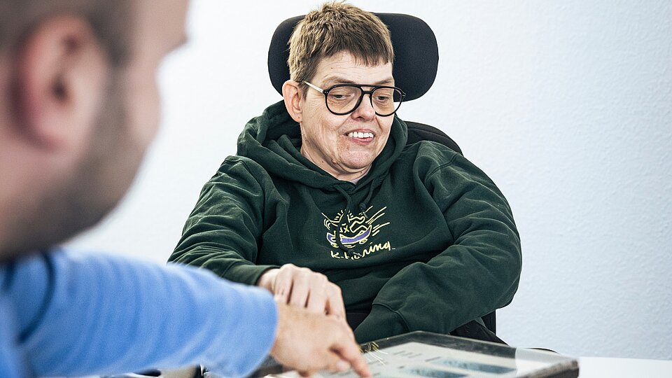 Eine Person mit Behinderung wird von einer anderen Person befragt und zeigt etwas auf einem Tablet.