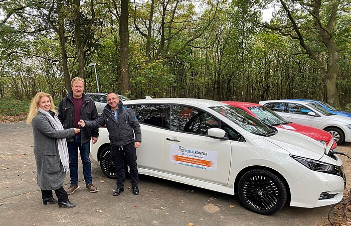 Drei lächelnde Mitarbeiter des Sozialkontors, zwei geben sich die Hand, im Hintergrund steht das erworbene E-Auto für Haus Eckel