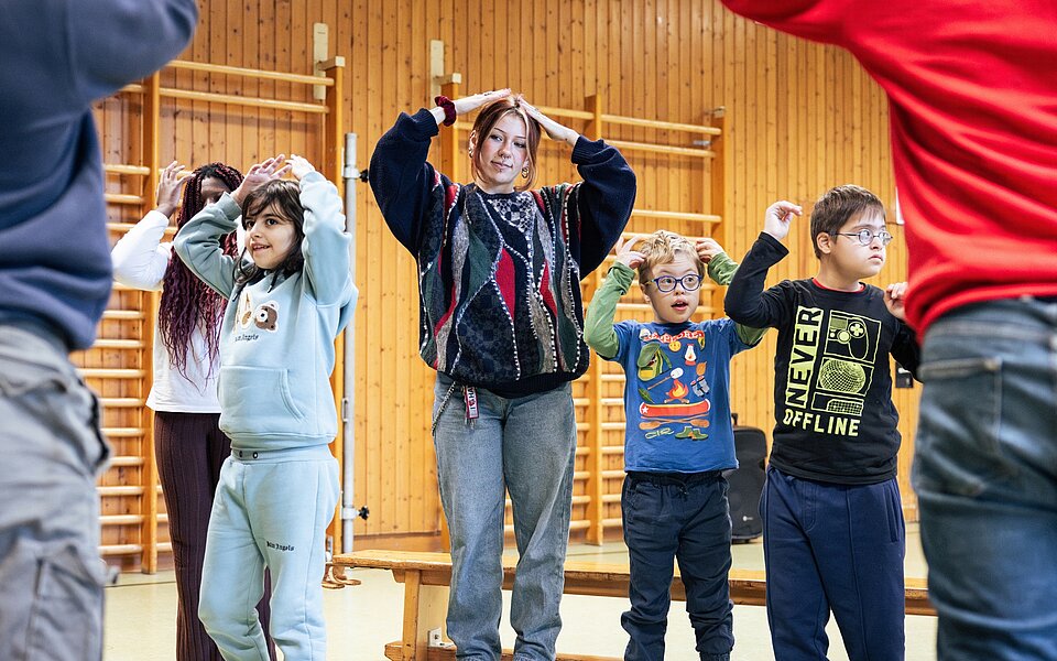 Kinder und Erwachsene machen gemeinsam eine Bewegungsübung in einer Turnhalle. Sie heben die Arme und halten ihre Hände über oder neben den Kopf.