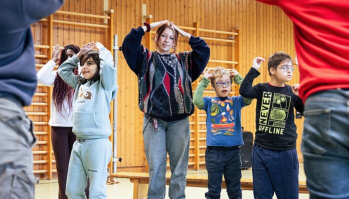 Kinder und Erwachsene machen gemeinsam eine Bewegungsübung in einer Turnhalle. Sie heben die Arme und halten ihre Hände über oder neben den Kopf.