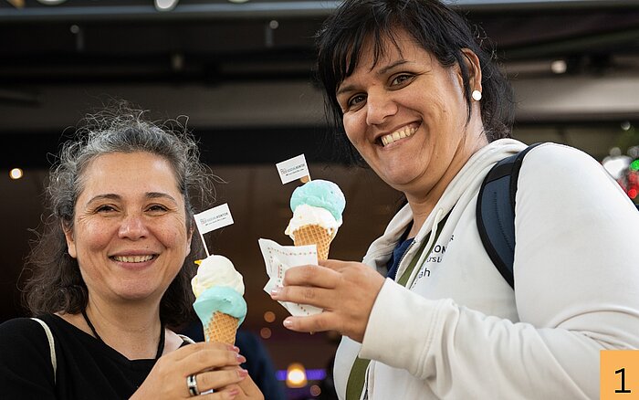 Ilkay Boran und Sarah Bartsch stehen lächelnd nebeneinander und halten Eiswaffeln mit blau-weißem Eis in den Händen.