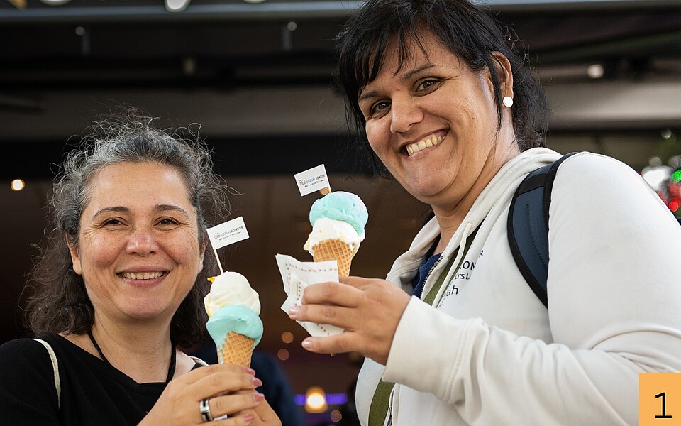 Ilkay Boran und Sarah Bartsch stehen lächelnd nebeneinander und halten Eiswaffeln mit blau-weißem Eis in den Händen.