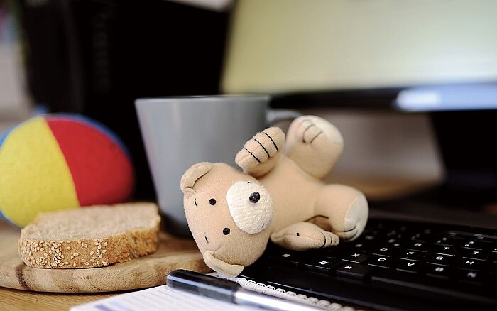 Ein Stofftier liegt vor einer Computertastatur, daneben ein Stück Brot, eine Kaffeetasse und ein bunter Ball auf einem Brett auf einem Schreibtisch.