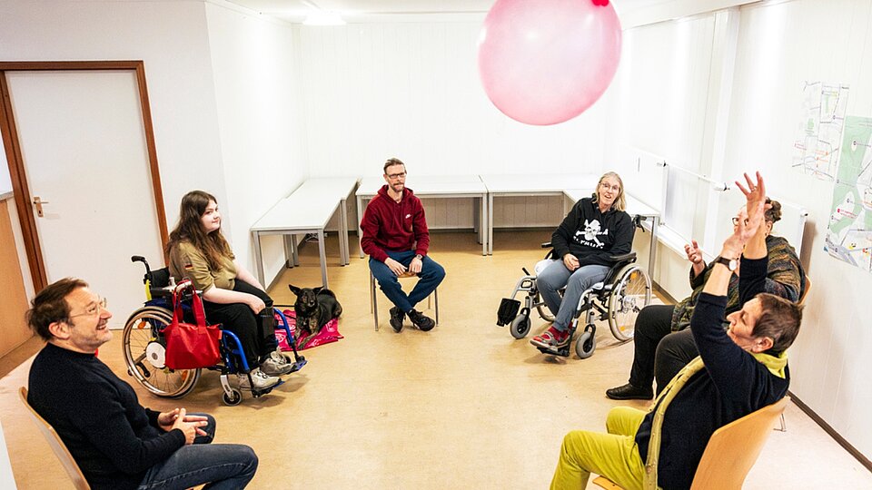 Bewegungsgruppe im Stuhlkreis wirft großen Ballon in die Luft; Rollstühle, Hund und Assistenzpersonen im Raum.