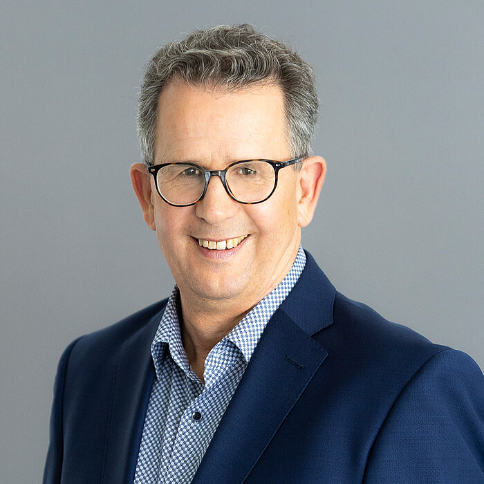 Gerd Müller, Geschäftsführung Finanzen und IT, ein Mann mit Brille und kurzem Haar, lächelt freundlich in die Kamera. Er trägt ein dunkles Jackett über einem rosa Hemd. Der Hintergrund ist dunkelgrau gestaltet.