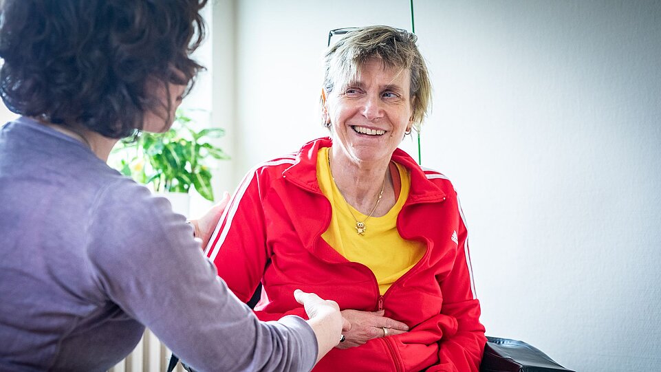 Eine Frau mit kurzen, blonden Haaren trägt eine rote Trainingsjacke über einem gelben T-Shirt und lächelt freundlich. Sie sitzt in einem Rollstuhl, während eine andere Frau mit dunklen, lockigen Haaren ihr die Hand reicht.