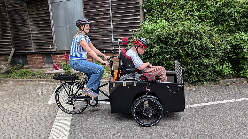 Zwei Personen fahren lächelnd auf dem E-Lastenrad des Hauses Beerboom auf einem asphaltierten Weg. Die vordere Person sitzt im Rollstuhl in der Transportbox, die speziell dafür ausgelegt ist. Die hintere Person fährt das Rad und tritt in die Pedale. Beide tragen Freizeitkleidung, und im Hintergrund sind einige grüne Büsche zu sehen.