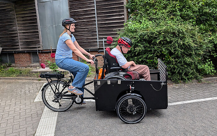 Zwei Personen fahren lächelnd auf dem E-Lastenrad des Hauses Beerboom auf einem asphaltierten Weg. Die vordere Person sitzt im Rollstuhl in der Transportbox, die speziell dafür ausgelegt ist. Die hintere Person fährt das Rad und tritt in die Pedale. Beide tragen Freizeitkleidung, und im Hintergrund sind einige grüne Büsche zu sehen.
