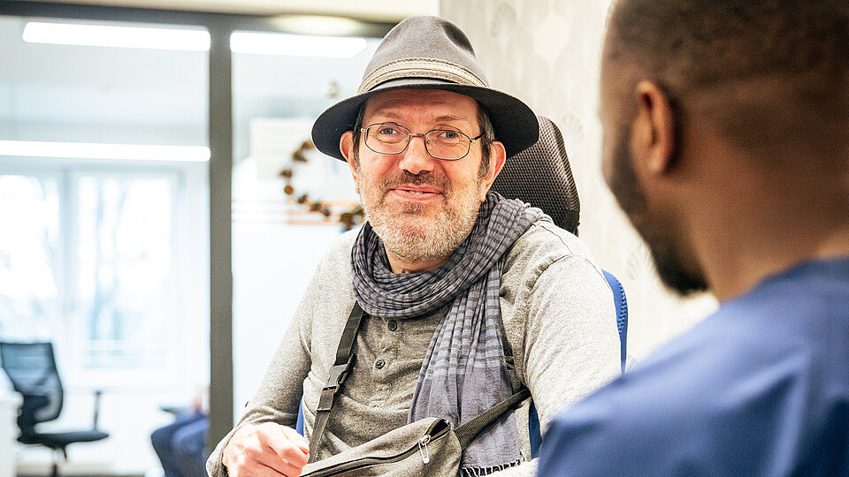 Ein älterer Mann im Rollstuhl trägt Hut und Schal und lächelt seinem Gegenüber freundlich zu. Der Gesprächspartner, ein Betreuer in blauer Kleidung, ist von hinten zu sehen.