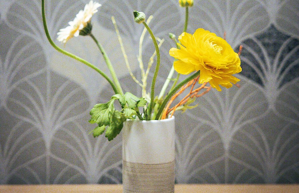 Ein schlichter Blumenstrauß mit gelben und weißen Blüten steht in einer weißen Vase auf einem Holztisch. Der Hintergrund zeigt eine Tapete mit dezentem Muster.