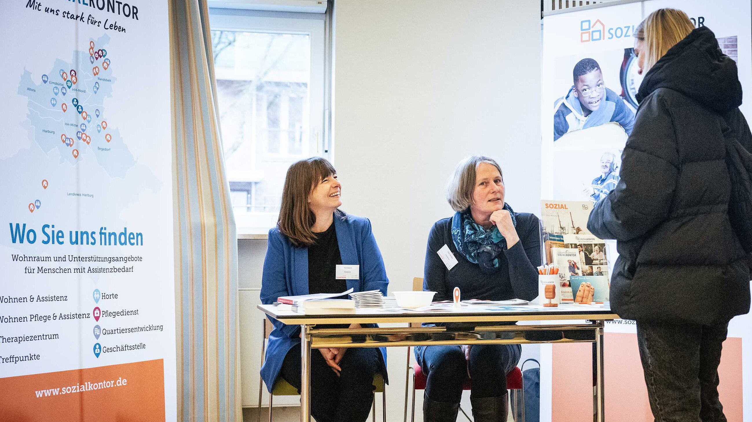 Zwei Frauen sitzen lächelnd an einem Infostand des Sozialkontors in einem Veranstaltungsraum. Eine Frau steht vor dem Tisch und spricht mit ihnen. Im Hintergrund sind Roll-Ups mit Infos zum Sozialkontor sichtbar. Auf dem Tisch liegen viele Materialien, Magazine und Flyer aus.