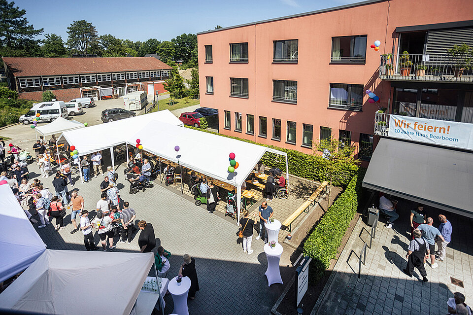 Blick von oben auf einen sonnigen Innenhof mit vielen Menschen bei der Jubiläumsfeier von Haus Beerboom. Es sind weiße Pavillons aufgebaut, unter denen Gäste stehen und sitzen. Rechts und links sind rot verklinkerte Gebäude mit mehreren Etagen vom Haus Beerboom zu sehen. Rechts hängt an einem Balkon ein Banner mit der Aufschrift: „Wir feiern! 10 Jahre Haus Beerboom“