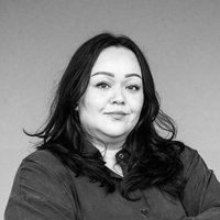 Eine Frau mit dunklen langen Haaren steht mit verschränkten Armen vor einem orangefarbenen Hintergrund. Sie schaut leicht zur Seite, mit einem neutralen Gesichtsausdruck.