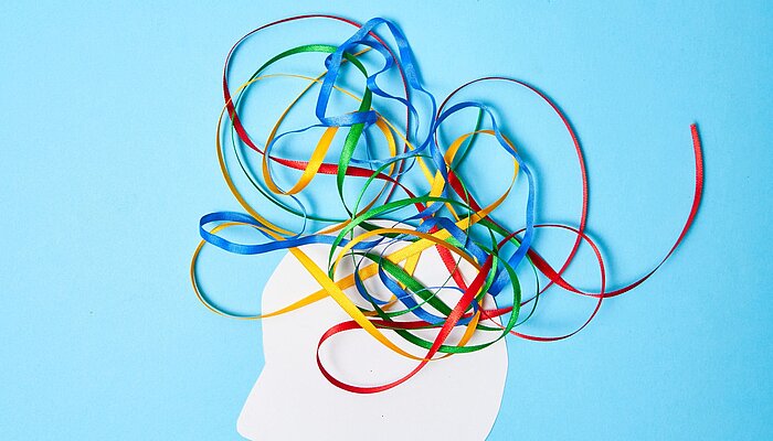 Silhouette eines menschlichen Kopfes in Weiß vor blauem Hintergrund, aus dem bunte Bänder in Rot, Blau, Grün und Gelb herauswirbeln, symbolisch für Gedanken, Kreativität und das Chaos im Kopf, passend zu ADS und ADHS.