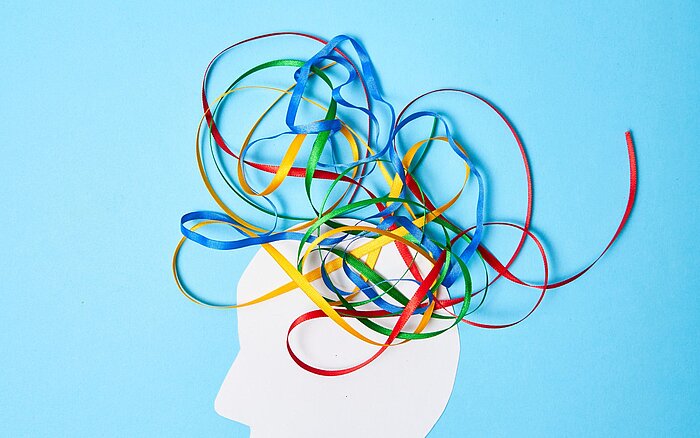 Silhouette eines menschlichen Kopfes in Weiß vor blauem Hintergrund, aus dem bunte Bänder in Rot, Blau, Grün und Gelb herauswirbeln, symbolisch für Gedanken, Kreativität und das Chaos im Kopf, passend zu ADS und ADHS.