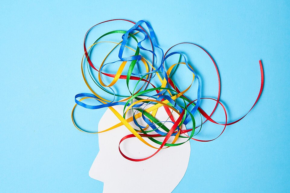 Silhouette eines menschlichen Kopfes in Weiß vor blauem Hintergrund, aus dem bunte Bänder in Rot, Blau, Grün und Gelb herauswirbeln, symbolisch für Gedanken, Kreativität und das Chaos im Kopf, passend zu ADS und ADHS.