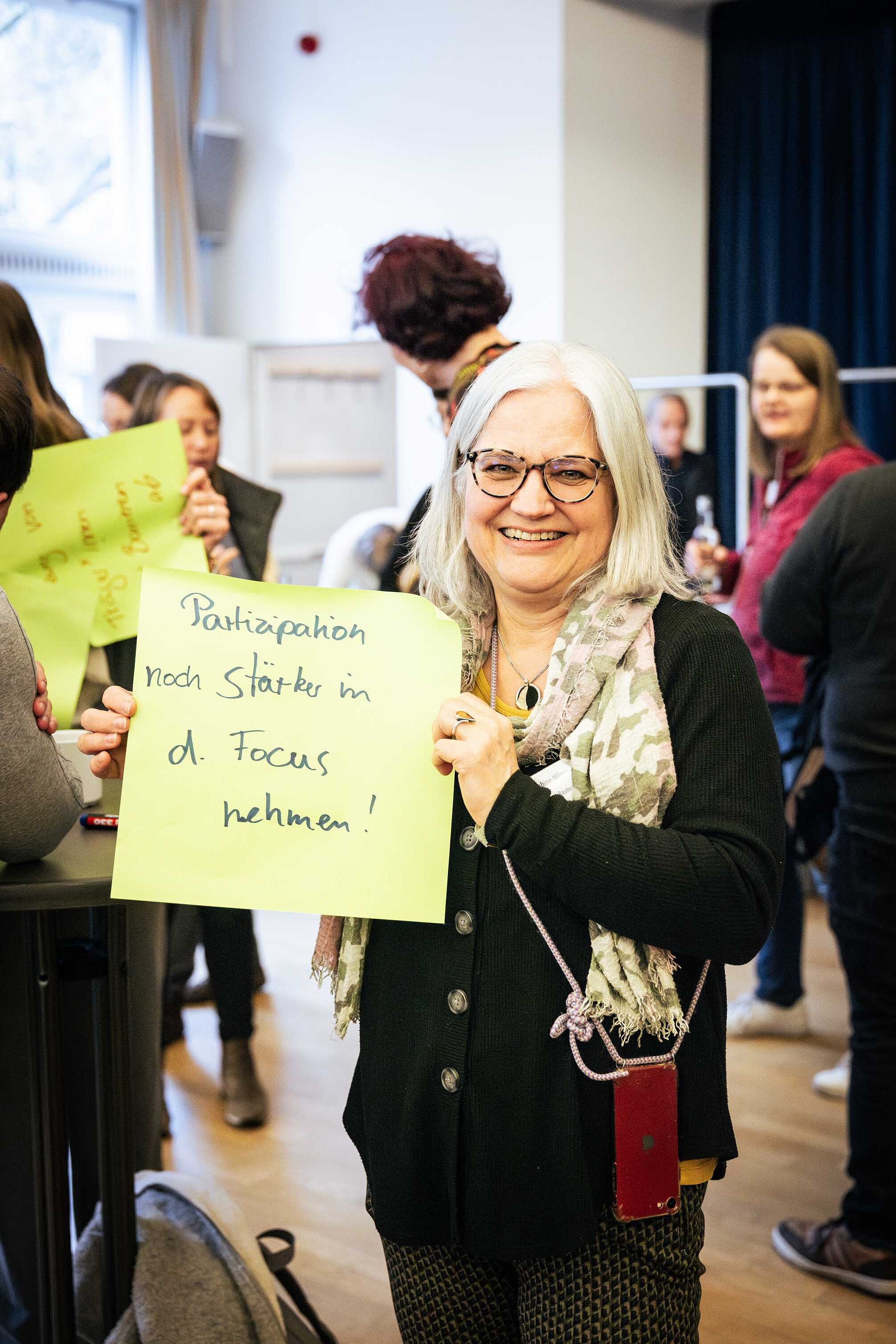 Eine Frau mit weißen Haaren hält einen großen gelben Zettel mit der Aufschrift „Partizipation noch stärker in den Fokus nehmen!“ in die Kamera und lächelt dabei. Im Hintergrund stehen weitere Teilnehmende der Veranstaltung. Einige davon halten ebenfalls große gelbe Zettel in der Hand.