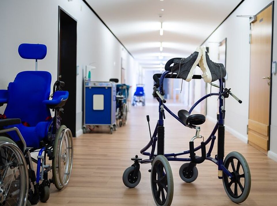 In einem langen Flur stehen verschiedene Hilfsmittel, darunter ein Rollstuhl und ein Gehtrainer mit vier Rädern. Auf dem Gehtrainer liegt ein Paar Schuhe.