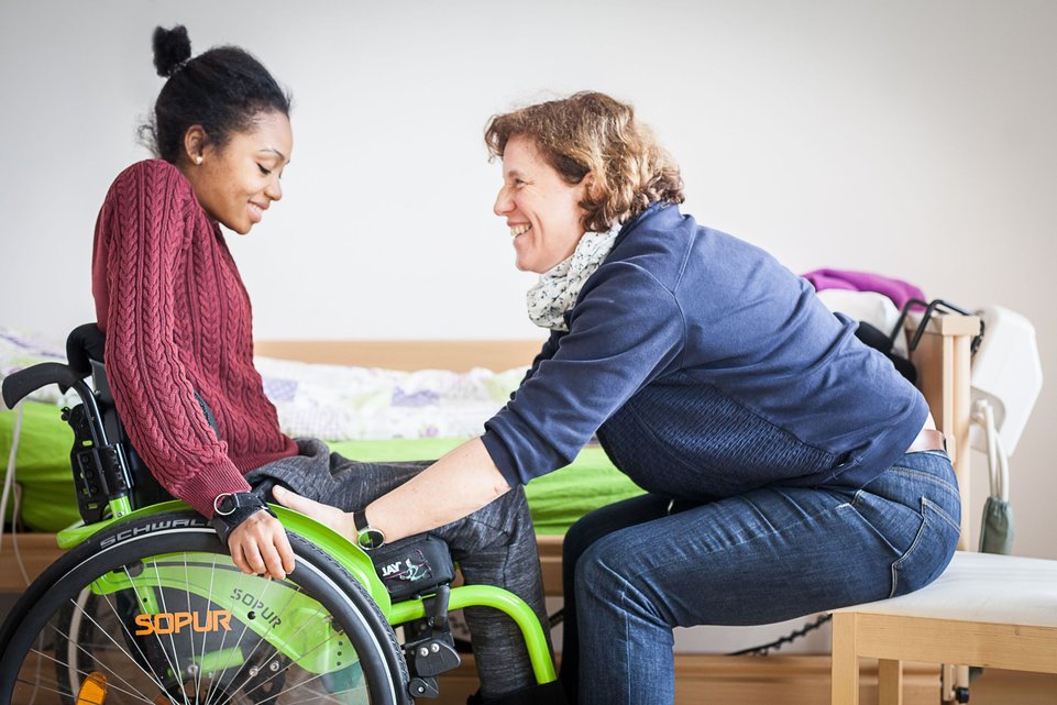 Zwei Frauen in einem Zimmer gegenübersitzend. Eine von ihnen sitzt im Rollstuhl.