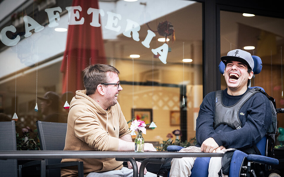 Vor einer Cafeteria sitzen zwei Männer an einem Tisch und lachen gemeinsam. Links sitzt ein Mann mit Brille und beigefarbenem Kapuzenpullover, rechts ein junger Mann im elektrischen Rollstuhl mit dunkelblauem Pulli, hellem Basecap und Armstützen. Beide wirken gelöst und fröhlich. Auf dem Tisch steht eine kleine Blumenvase. Im Hintergrund ist durch das Fenster der Innenbereich der Cafeteria zu sehen.