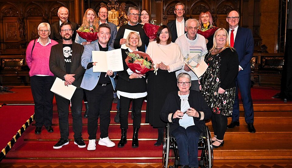 Eine Gruppe von Personen, die in einem formellen Rahmen für ein Foto posieren, wobei einige Urkunden und Blumensträuße in der Hand halten, bei einer Preisverleihung zum 
