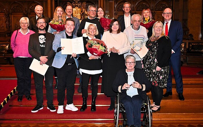 Eine Gruppe von Personen, die in einem formellen Rahmen für ein Foto posieren, wobei einige Urkunden und Blumensträuße in der Hand halten, bei einer Preisverleihung zum