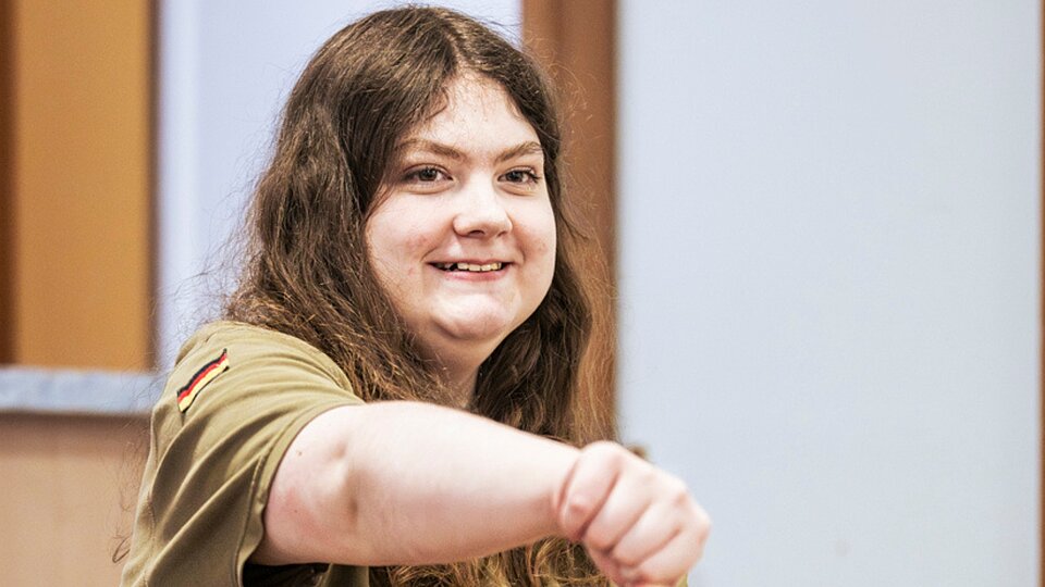 Junge Rollstuhlnutzerin lächelt und streckt den Arm nach vorn in einer Übung.