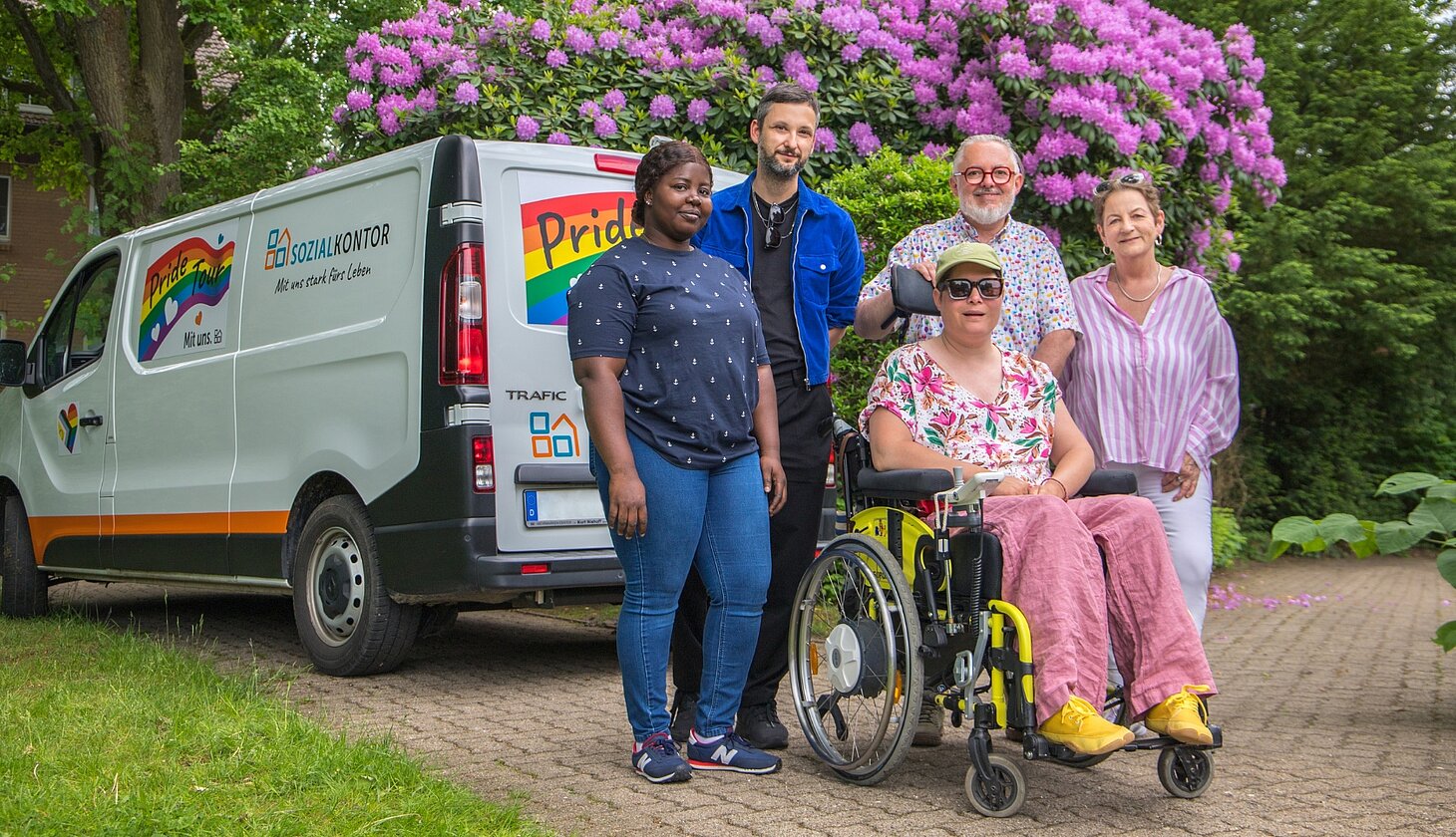 Menschen mit und ohne Behinderung stehen vor einem Pride-Tour-Fahrzeug des Sozialkontors und kommen miteinander ins Gespräch. Das Fahrzeug ist mit Regenbogenfarben gestaltet und wirbt für Vielfalt und Akzeptanz.