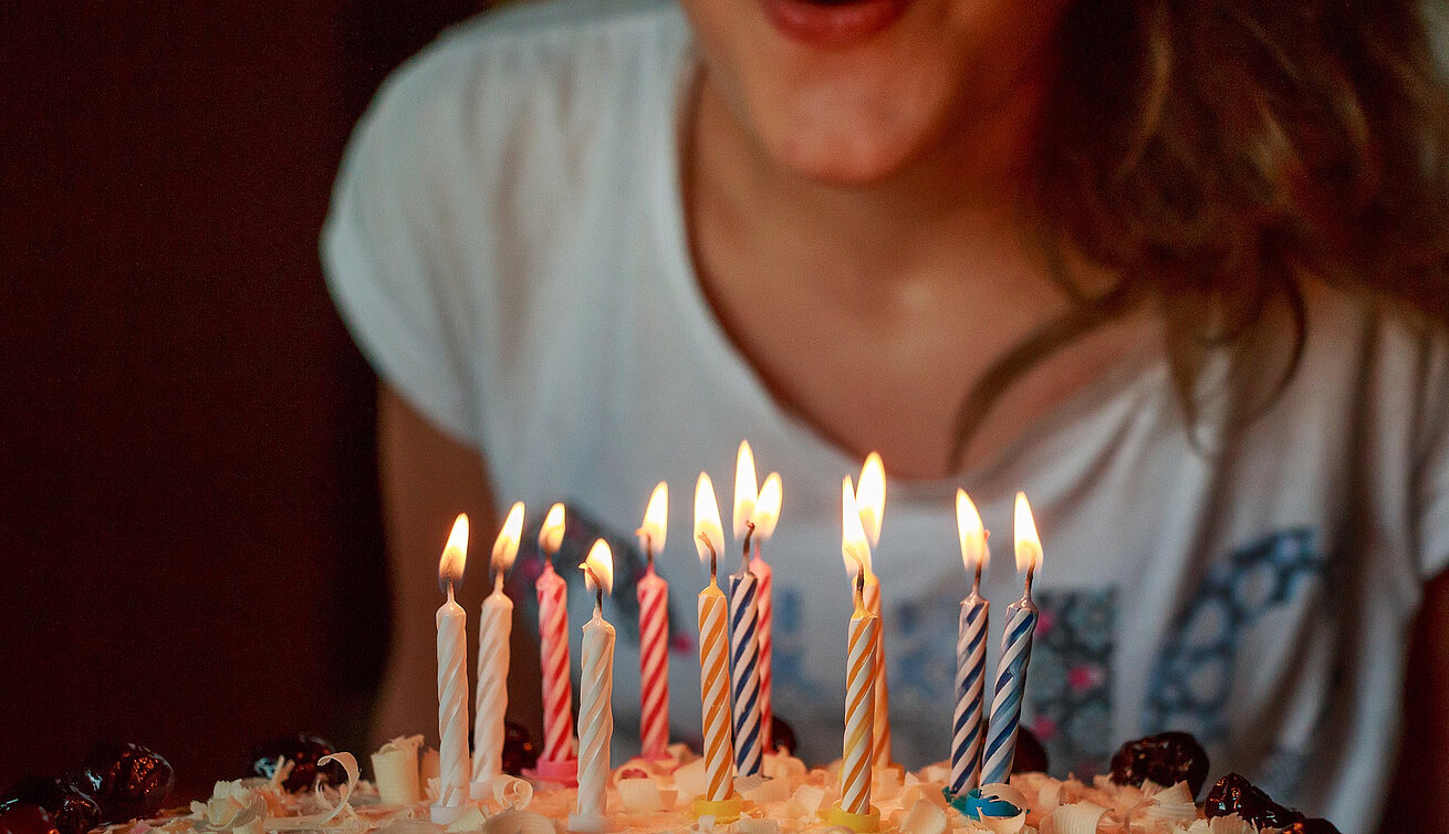 Im Vordergrund steht ein Kuchen mit mehreren brennenden Geburtstagskerzen. Dahinter beugt sich eine Person in einem weißen T-Shirt über den Kuchen.