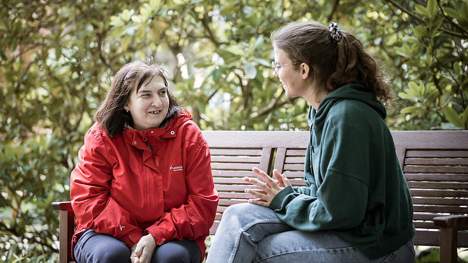 Zwei Frauen sitzen auf einer Holzbank im Grünen und unterhalten sich angeregt. Eine trägt eine rote Jacke, die andere eine grüne. Beide wirken entspannt und zugewandt.