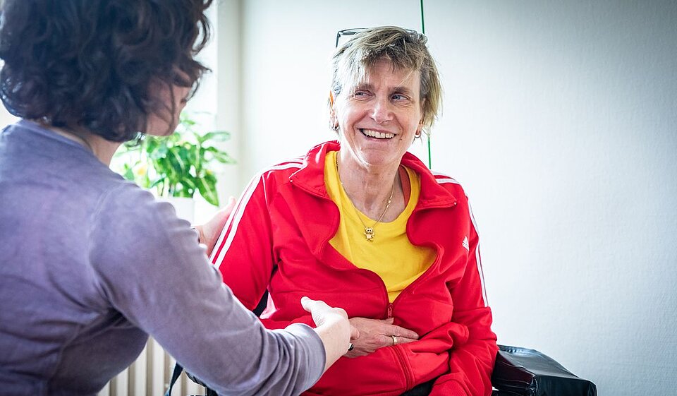 Zwei Personen sitzen sich gegenüber und halten sich an den Händen. Eine Person trägt eine rote Jacke und ein gelbes T-Shirt, die andere ist seitlich im Vordergrund zu sehen.