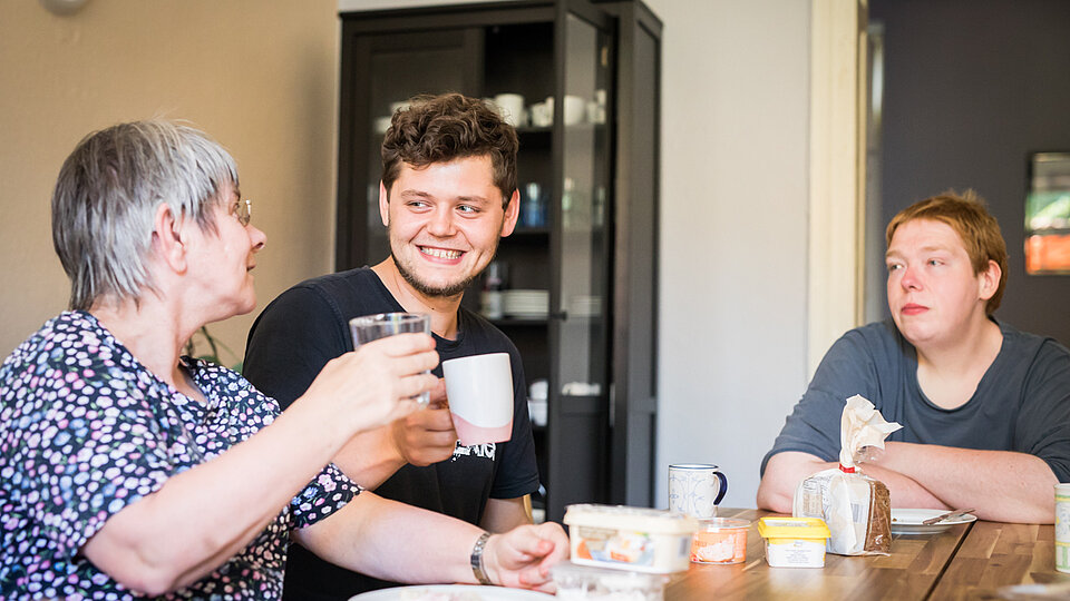 Drei Personen sitzen an einem Esstisch und stoßen mit Getränken fröhlich an. Auf dem Tisch stehen Frühstücksutensilien.