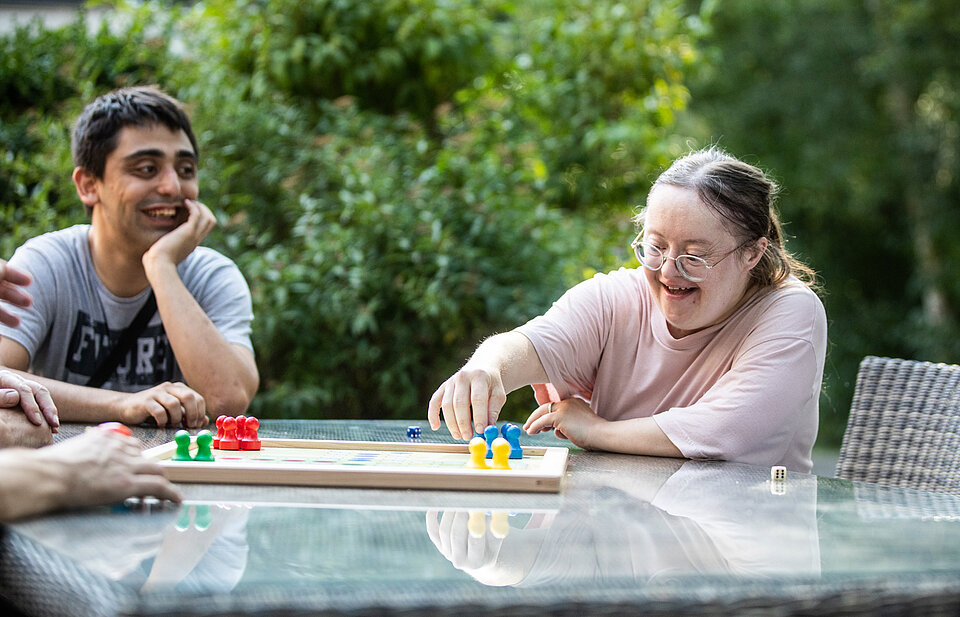 Zwei junge Erwachsene spielen lachend ein Brettspiel im Freien an einem Glastisch. Eine Frau mit Down-Syndrom greift fröhlich nach einer Spielfigur, ein junger Mann schaut sie lächelnd an.