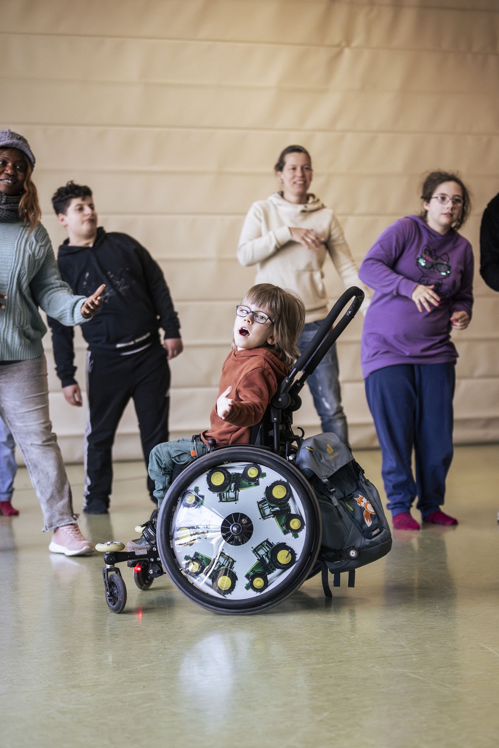 Ein Kind im Rollstuhl mit auffälligen Traktor-Motiven auf den Rädern tanzt mit erhobenem Arm inmitten einer Gruppe von Kindern und Erwachsenen.
