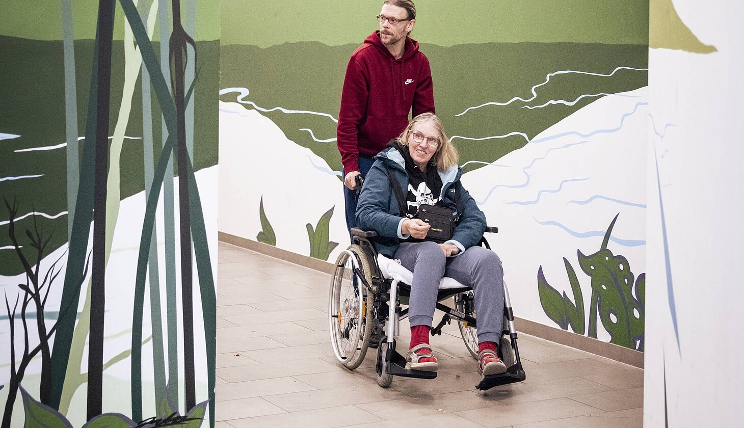 Ein Mann schiebt eine Frau im Rollstuhl durch einen hellen Flur mit Wandmalereien von Bergen und Natur.