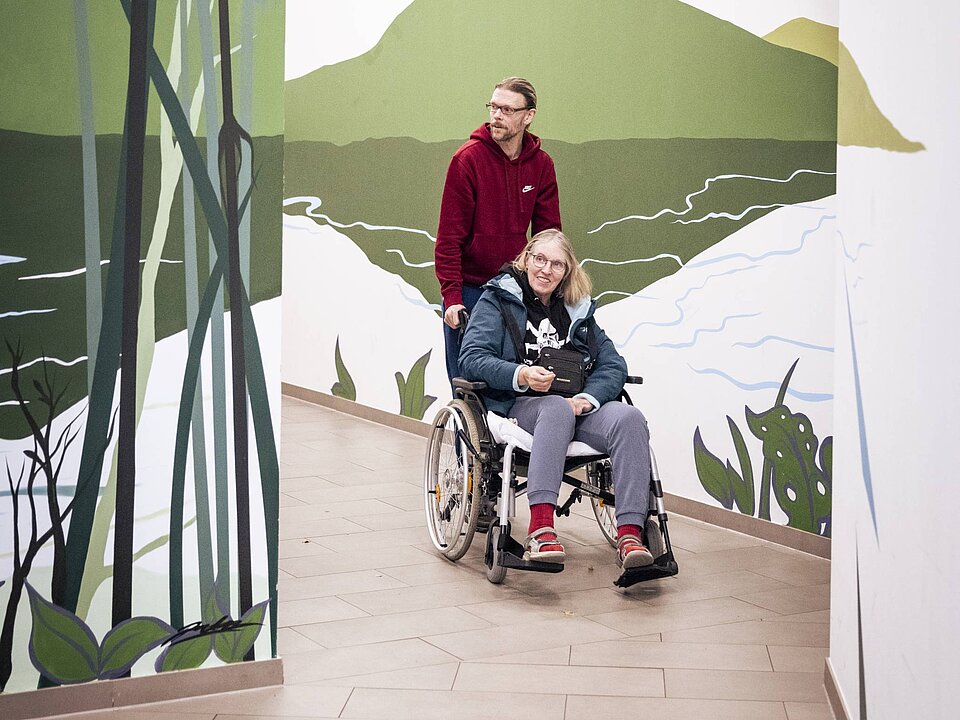 Ein Mann schiebt eine Frau im Rollstuhl durch einen hellen Flur mit Wandmalereien von Bergen und Natur.