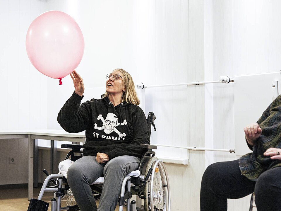 Eine Frau im Rollstuhl sitzt in einem Zimmer und stößt einen großen rosa Luftballon durch die Luft. Sie trägt einen schwarzen Kapuzenpullover mit einem Totenkopf und der Aufschrift 
