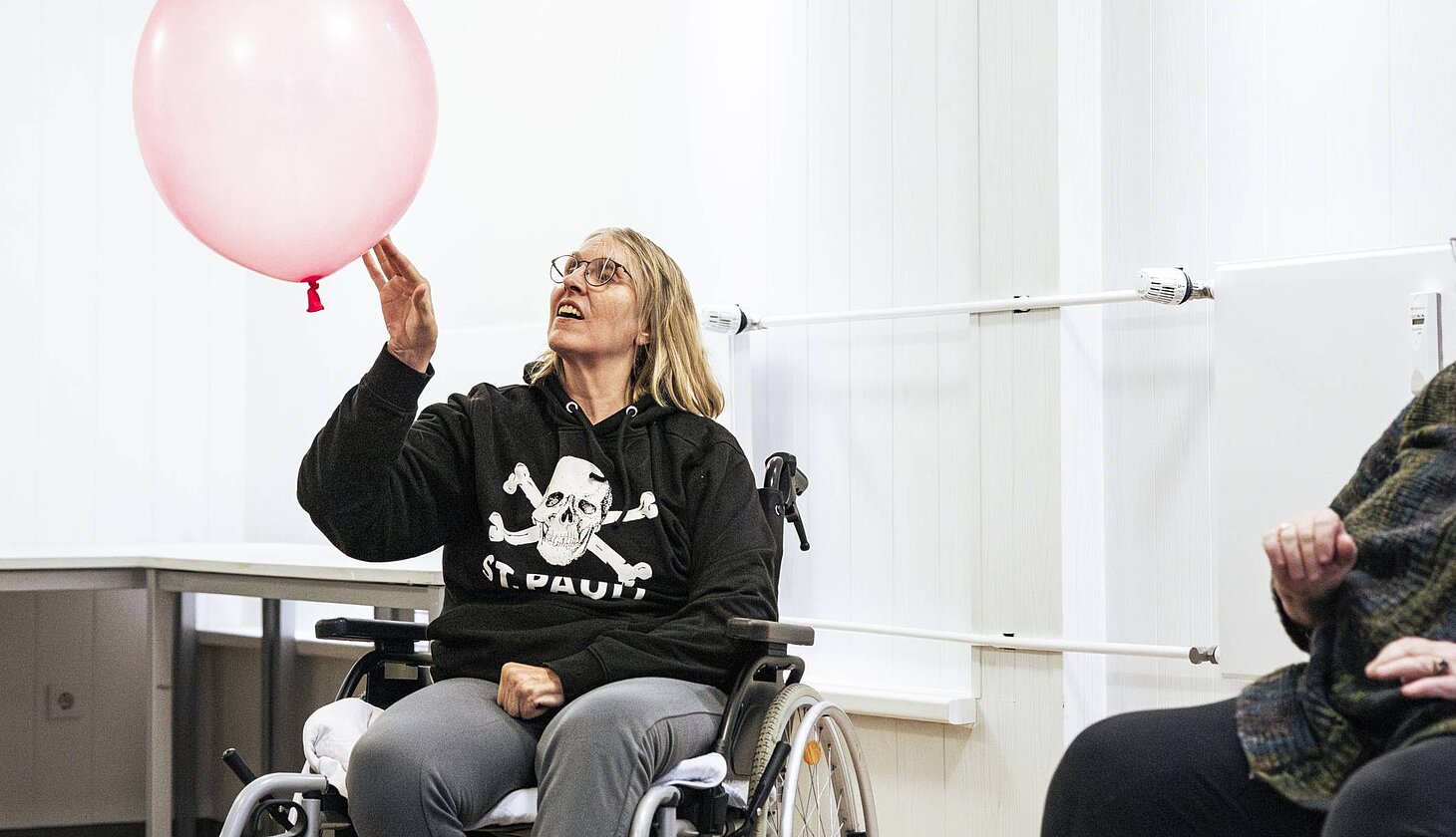 Eine Frau im Rollstuhl sitzt in einem Zimmer und stößt einen großen rosa Luftballon durch die Luft. Sie trägt einen schwarzen Kapuzenpullover mit einem Totenkopf und der Aufschrift 