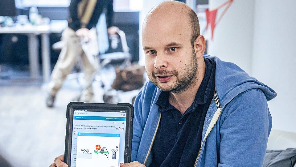 Niklas Luy zeigt ein Tablet mit Symbolen aus einer Befragungs-App. Die Bilder helfen dabei, Fragen zur Assistenz verständlicher zu machen.