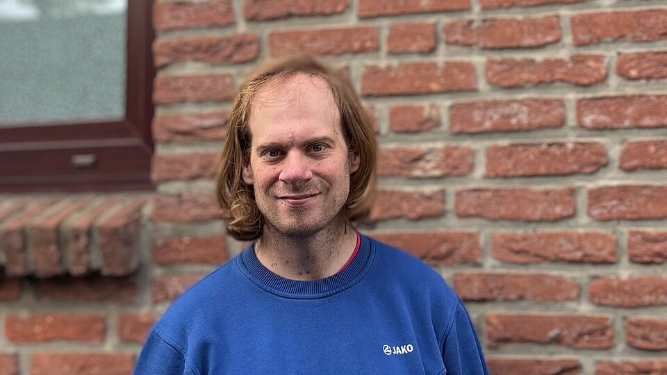 Ein Mann in einem blauen Pullover vor einer Backstein-Hausmauer.