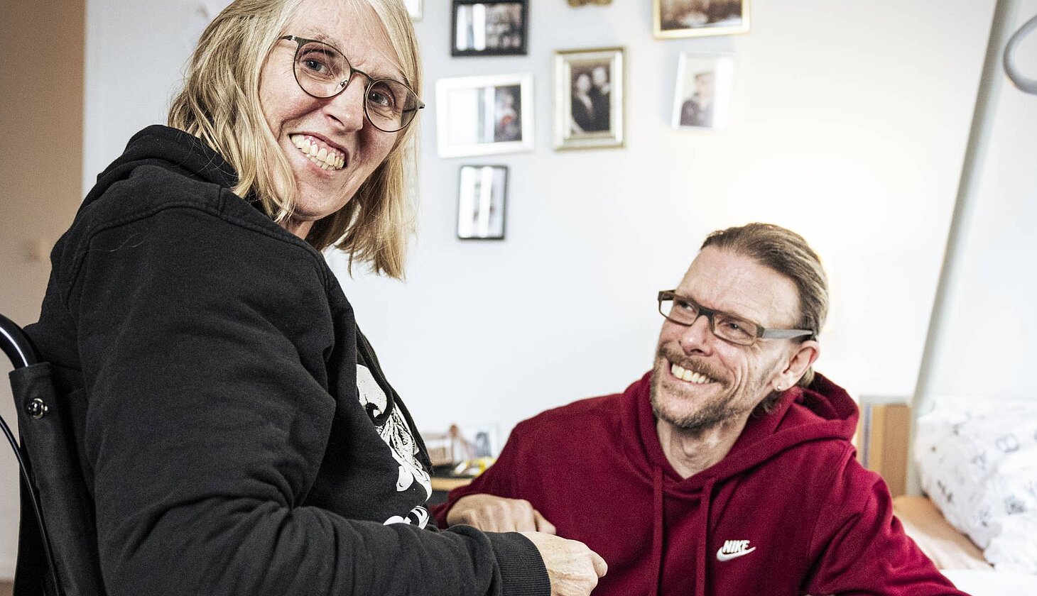 Eine Frau im Rollstuhl sitzt in einer Wohnung und lächelt, während ein Mann neben ihr sitzt und sie anschaut.