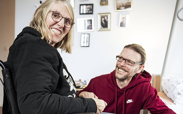 Eine Frau im Rollstuhl sitzt in einer Wohnung und lächelt, während ein Mann neben ihr sitzt und sie anschaut.