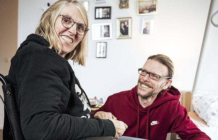Eine Frau im Rollstuhl sitzt in einer Wohnung und lächelt, während ein Mann neben ihr sitzt und sie anschaut.