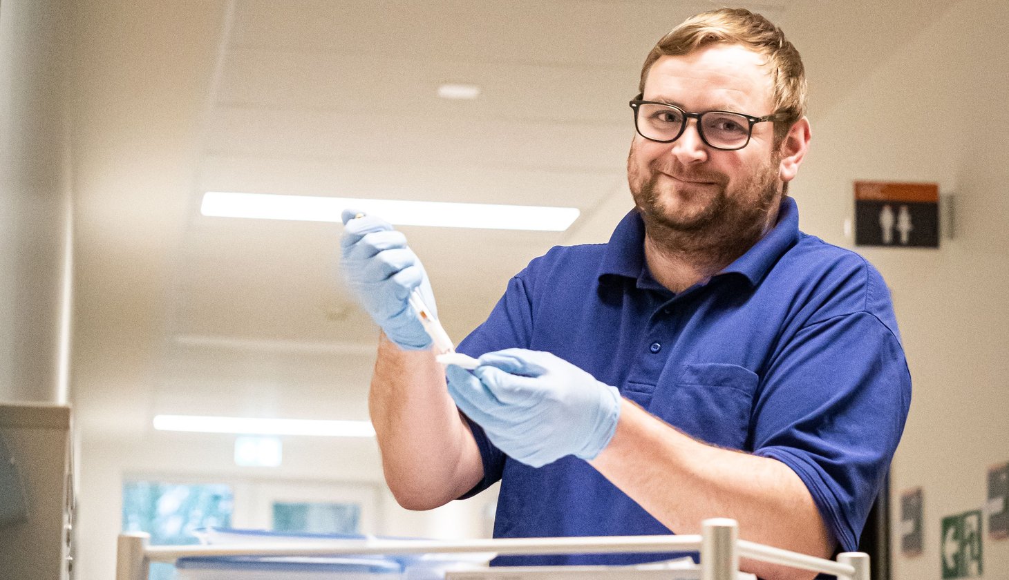 Eine lächelnde Pflegefachkraft mit Brille, blauen Gummihandschuhen und einem marineblauen Polohemd steht in einem Krankenhaus an einem Tisch und bereitet eine Spritze vor.