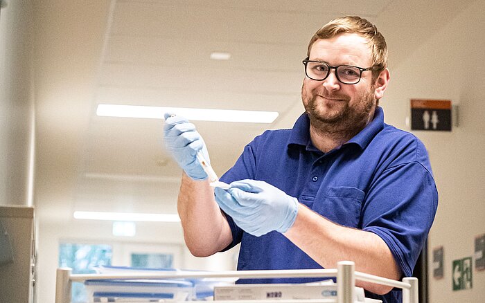 Eine lächelnde Pflegefachkraft mit Brille, blauen Gummihandschuhen und einem marineblauen Polohemd steht in einem Krankenhaus an einem Tisch und bereitet eine Spritze vor.