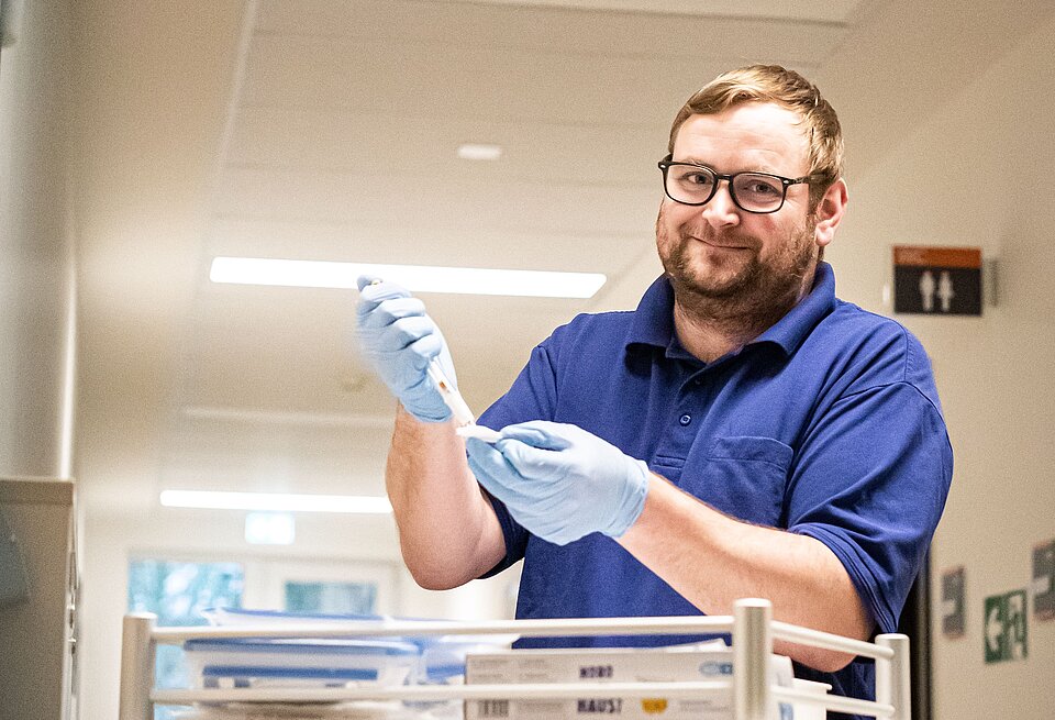 Eine lächelnde Pflegefachkraft mit Brille, blauen Gummihandschuhen und einem marineblauen Polohemd steht in einem Krankenhaus an einem Tisch und bereitet eine Spritze vor.