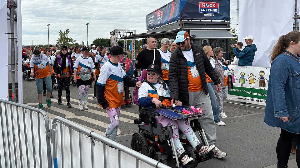 Menschen mit und ohne Behinderung nehmen gemeinsam am HafenCity Run teil. Im Vordergrund schiebt eine Person eine Teilnehmerin im Rollstuhl, beide tragen bunte Sozialkontor-Shirts und lächeln. Weitere Personen gehen im Hintergrund über die Ziellinie. Die Stimmung ist lebendig und inklusiv, begleitet von Bannern und Zuschauenden am Rand.