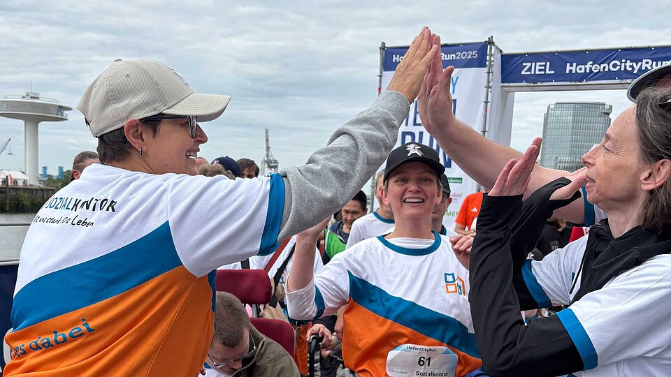 Teilnehmende des HafenCity Run geben sich mit strahlenden Gesichtern ein High-Five im Zielbereich. Sie tragen Sozialkontor-Laufshirts. Im Hintergrund: der Hafen und das ZIEL-Banner.