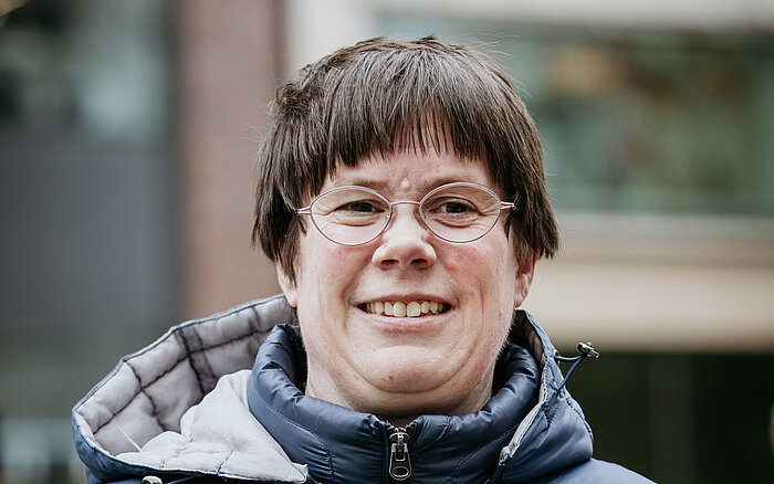 Katrin Schönfeld bei einer Veranstaltung im Freien. Sie trägt eine wattierte, dunkelblaue Jacke mit Kapuze und eine randlose Brille. Sie lächelt in die Kamera und hält ein Schild oder Papier in der Hand. Der Hintergrund ist unscharf, es sind Gebäude und Menschen im öffentlichen Raum zu erkennen.